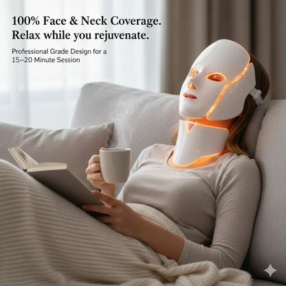 Person wearing a LED face and neck mask while reading a book and holding a mug on a couch.